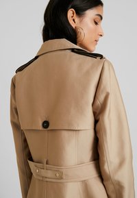 Beige trench coat with a structured silhouette, featuring a large collar, a button detail at the back, and a waist belt for adjustment.