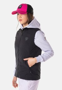 Schwarze Weste mit Reißverschluss, darunter ein Kapuzenpullover und eine pinkfarbene Netz-Cap. Auffällige Details: Logo auf der Brust und Farbkontrast im Stoff.