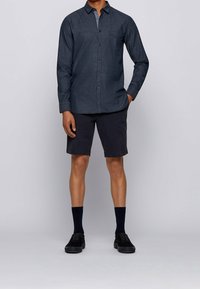 Mörkblå skjorta med knapp-up med en subtil textur, långa ärmar och en ficka, kombinerad med mörka shorts och svarta skor och strumpor.