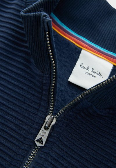 Giacca a zip blu navy con texture a coste, colletto a righe colorate, zip argentata con tirante con marchio e etichetta bianca che recita "Paul Smith JUNIOR."