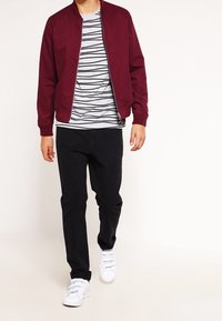 Bordeaux bomberjacka över en grå och vit randig skjorta, matchad med svarta byxor och vita sneakers. Enkel design med ribbade  ärmslut.