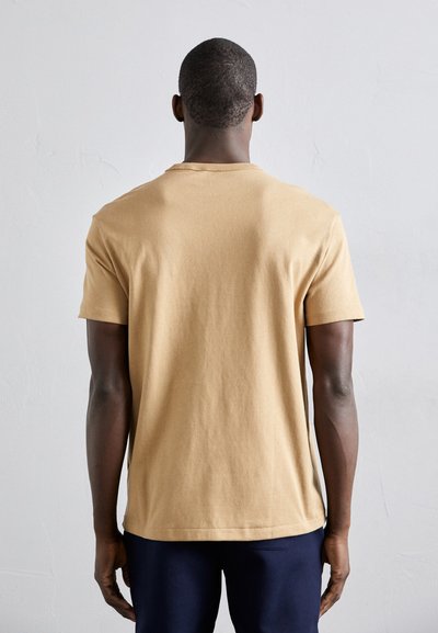 Beige cotton t-shirt with short sleeves, rounded neckline, and a relaxed fit, worn with dark navy pants. Simple, unadorned design.
