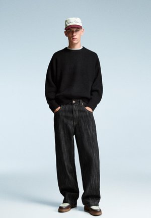 Black knit sweater, loose-fit dark denim jeans, and brown-and-white shoes. Model wears a light-colored cap with text. Simple background.