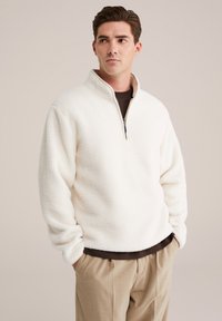 Pullover in pile bianco con colletto a mezzo zip, caratterizzato da una consistenza morbida. Indossato sopra una camicia scura con pantaloni color sabbia.