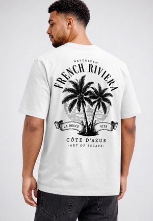 Mann trägt ein weißes T-Shirt mit schwarzem Palmen-Design und dem Text French Riviera, La Dolce Vita, Côte d'Azur, Art of Escape.
