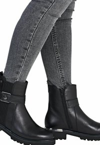 Botines de cuero negro hasta el tobillo con un panel texturizado, cremallera lateral y detalle de hebilla. Cuentan con una suela de goma resistente con agarre.