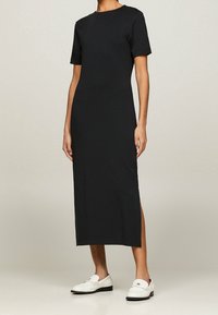 Zwarte midi-jurk met korte mouwen, gemaakt van jersey-materiaal, met een ronde halslijn en zijsplitten. Gekozen in combinatie met witte instappers.