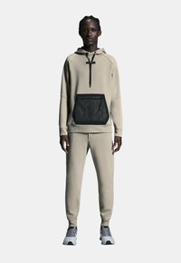 Beige hoodie en joggingsbroek gemaakt van zachte stof, met een zwart gaasvak aan de voorkant. Gestyled met grijze sportschoenen.
