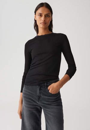 Una donna con lunghi capelli scuri indossa una maglietta nera a costine a maniche lunghe e jeans neri sbiaditi, posando con una mano in tasca su uno sfondo semplice.