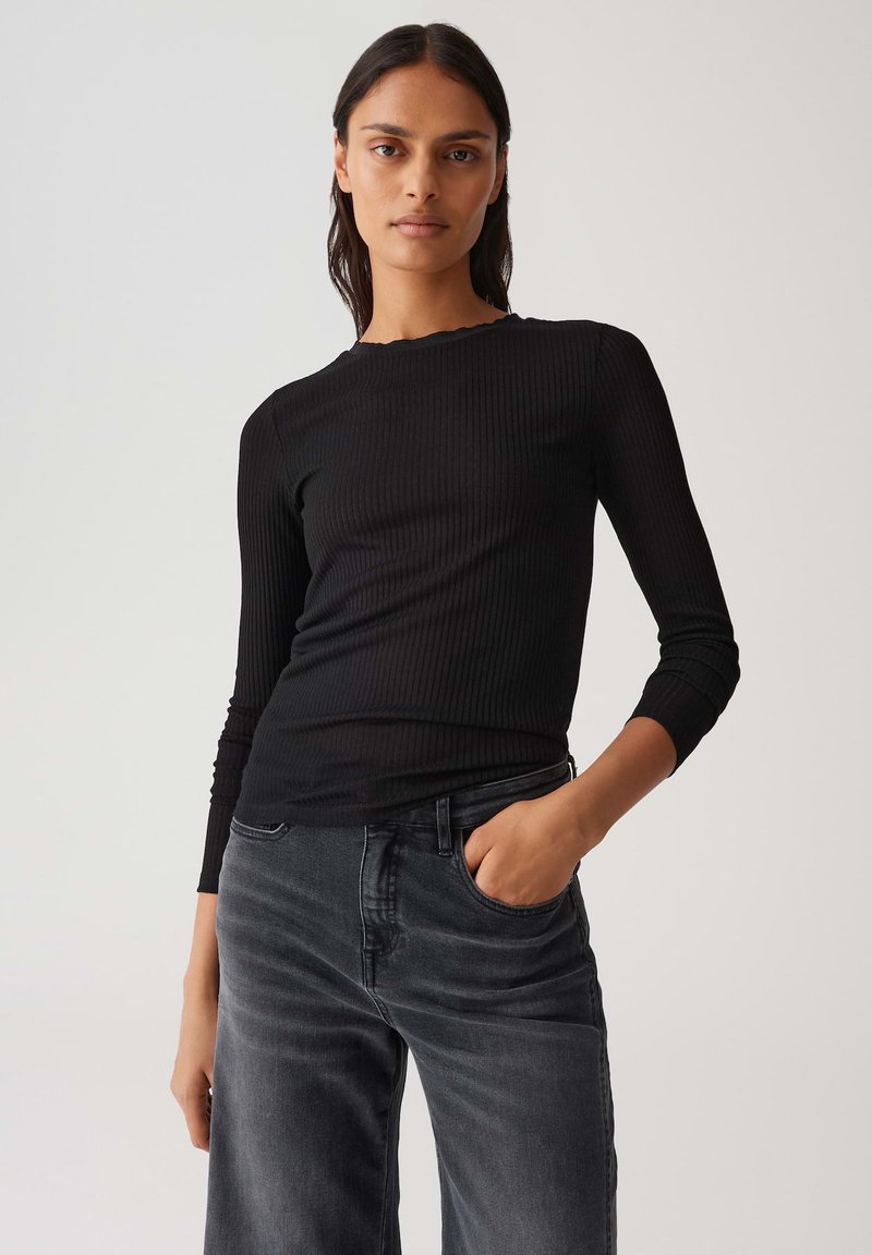 Femme aux longs cheveux noirs portant un top noir à manches longues côtelé et un jean noir délavé, posant avec une main dans sa poche contre un fond uni.