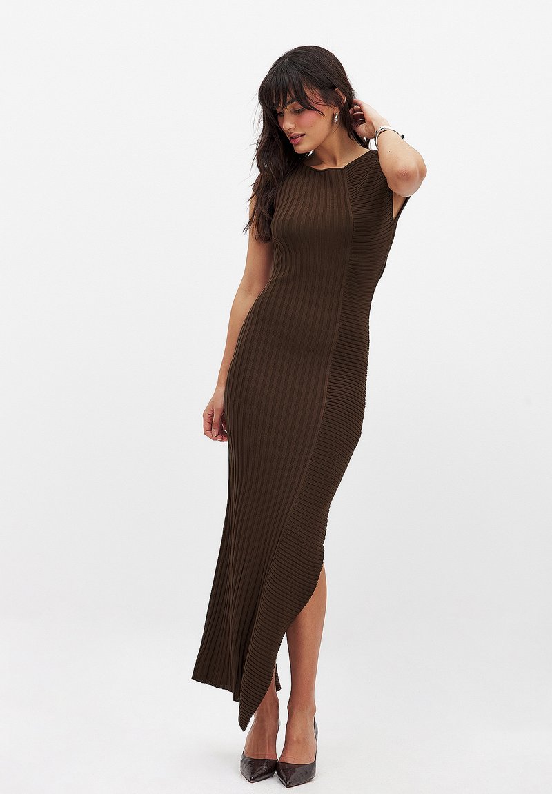 Robe marron à côtes avec une fente haute, sans manches, silhouette ajustée et texture lisse. Portée avec des chaussures à bout pointu.