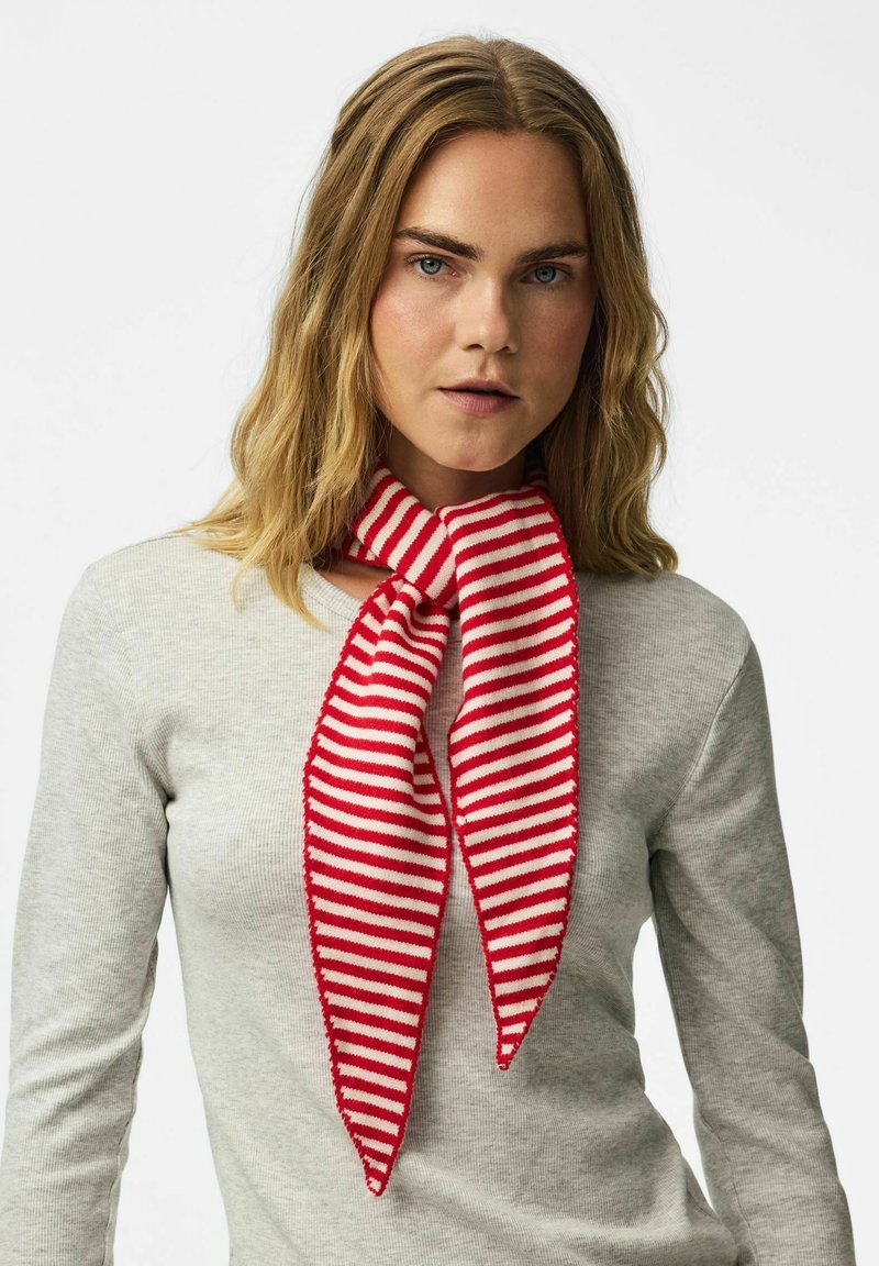 Femme aux cheveux blonds portant un haut gris clair à manches longues et une écharpe rouge et blanche rayée nouée autour du cou, sur un fond uni.