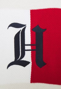 Lettre gothique noire "H" brodée sur un tissu blanc tricoté avec un fond de rayure verticale rouge tricotée.