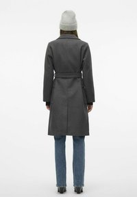 Cappotto di lana grigio con vita regolabile, maniche lunghe e colletto classico. Abbinato a jeans blu e a un berretto lavorato a maglia grigio chiaro.