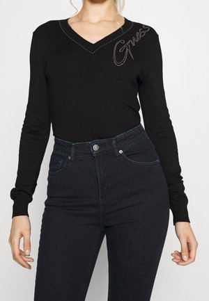 Femme portant un top noir manches longues à col V avec "Guess" en strass sur l'épaule et un jean noir taille haute.