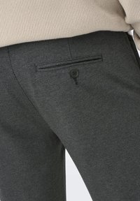Grå herringbone-byxor med en knappad bakficka och en texturerad yta, bärs med en beige stickad topp delvis instoppad.