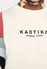 Felpa color crema con pannelli sulle spalle multicolori in rosso, verde, azzurro e nero; presenta un logo ricamato "KAOTIKO" e "SINCE 1999."