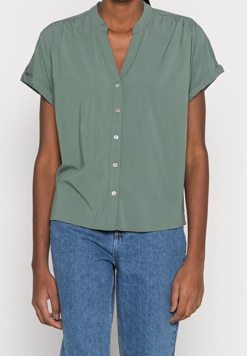 Persona che indossa una blusa verde salvia a maniche corte con bottoni abbinata a jeans blu a vita alta, su uno sfondo semplice.