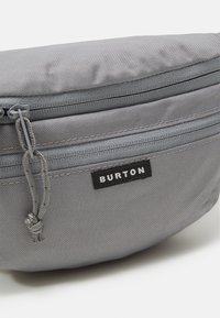 Szürke szövet zsák, texturált, tartós felülettel. A tetején cipzárral és egy fekete "BURTON" feliratú címkével rendelkezik. Kompakt és letisztult design.