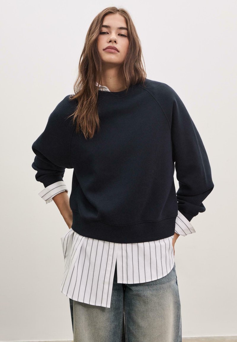 Felpa blu navy con maniche raglan, polsini e orlo a coste. Abbinata a una camicia bianca a righe indossata con jeans a gamba larga di un blu chiaro.