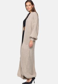 Long, beige knitted cardigan with a loose fit, dropped shoulders, and a front pocket. Worn over a black crop top and paired with black heels.