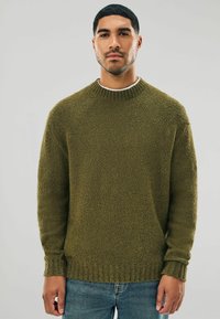 Olive green sweater in a textured knit, featuring a crew neck and ribbed cuffs and hem. Worn over a white shirt, paired with denim jeans.