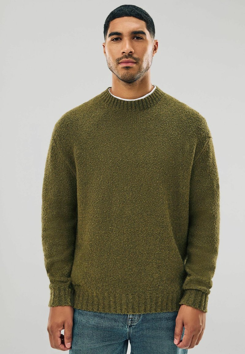 Olive green sweater in a textured knit, featuring a crew neck and ribbed cuffs and hem. Worn over a white shirt, paired with denim jeans.