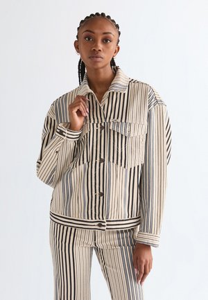 Žena s vrkočmi, oblečená v béžovo-čiernom prúžkovanom saku a nohaviciach, stojaca pred bielym pozadím.