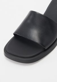 Mules noires en cuir synthétique lisse, avec une large bande supérieure et une semelle plate rembourrée de forme rectangulaire.