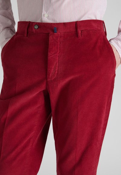 Pantalones de pana rojos con estrías verticales, que cuentan con un bolsillo lateral y un cierre delantero con botón. Accentuados por una pequeña etiqueta azul.