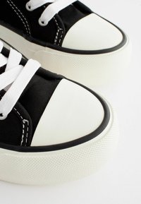 Schwarze Leinensneaker mit weißen Gummi-Spitzen, strukturierten weißen Sohlen und schwarzen Akzenten. Die Schnürsenkel sind weiß und nähtechnische Details sind sichtbar.