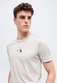 Νεαρός άνδρας με κοντά σγουρά μαλλιά που φοράει ανοιχτό μπεζ μπλουζάκι BOSS και ασημένιο κολιέ, κοιτάζοντας προς τα πλάγια με φόντο απλό.