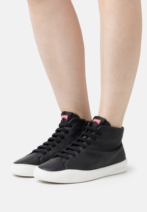 Baskets montantes noires avec semelles blanches portées sur jambes nues, avec lacets noirs et une étiquette rouge sur la languette.