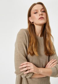 Beżowy sweter w prążki z okrągłym dekoltem i mankietami. Modelka pozuje z założonymi rękami, mając długie proste włosy i kolczyki w kształcie obręczy.