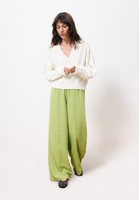 Weißer Strickpullover mit tiefem V-Ausschnitt und gerippten Bündchen, kombiniert mit fließenden, weit geschnittenen hellgrünen Hosen und schwarzen gemusterten Schuhen.