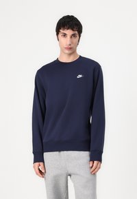 Marineblauwe Nike-sweater met een ronde hals, lange mouwen en logo op de linkerkant van de borst; gecombineerd met lichtgrijze sweatpants.