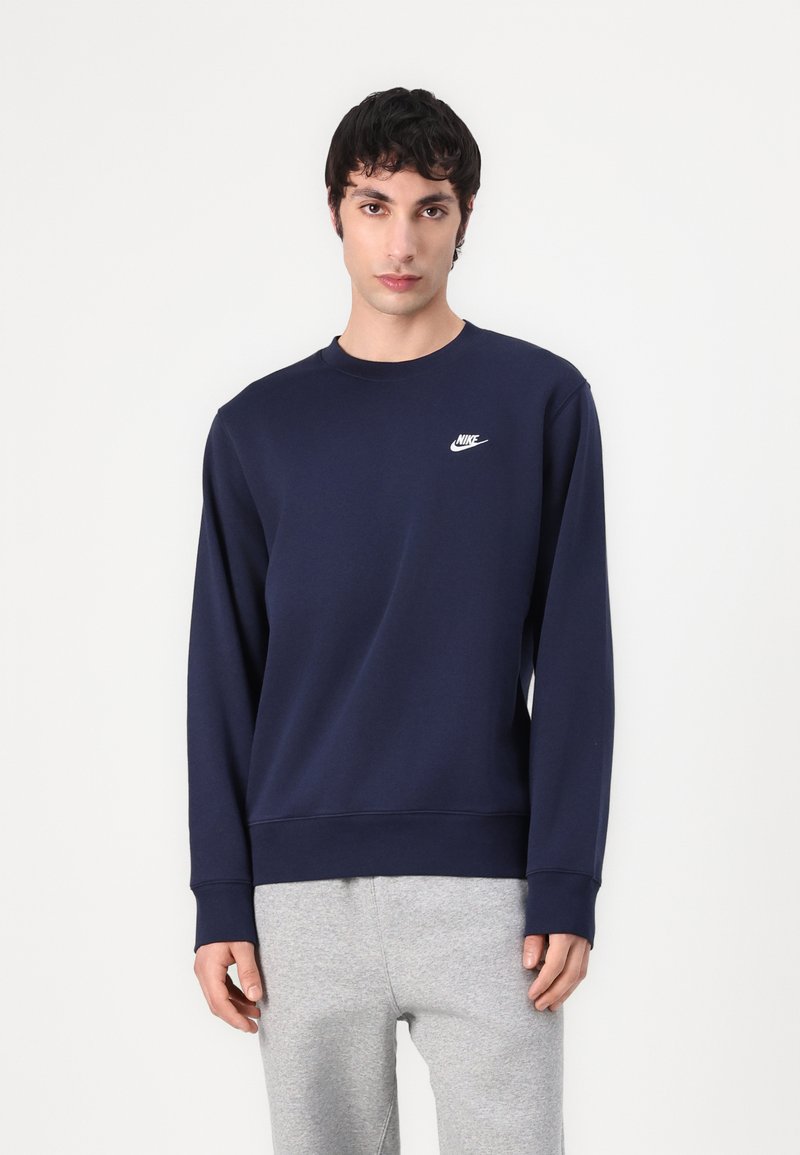 Marineblauwe Nike-sweater met een ronde hals, lange mouwen en logo op de linkerkant van de borst; gecombineerd met lichtgrijze sweatpants.