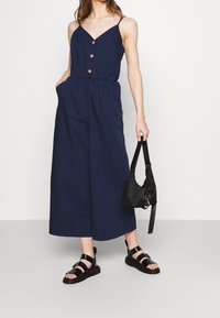 Marineblauer Overall mit V-Ausschnitt, verstellbaren Spaghetti-Trägern, Holzknöpfen und elastischem Bund. Kombiniert mit schwarzen Riemchensandalen und Tasche.