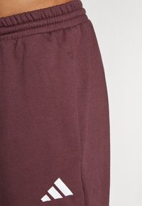 Burgundijas sporta bikses ar elastīgu jostasvietu. Iespējas ar baltām trīs svītru logo detaļām pie apakšējās malās. Mīksta auduma tekstūra, pieguļošs dizains.