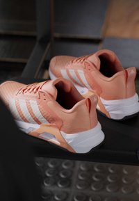 Двойка коралово розови маратонки Adidas с бели подметки и три бели ивици, поставени на черна пейка в зала за тренировки или съблекалня.