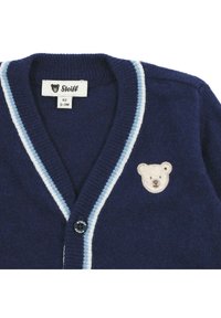 Marineblauwe cardigan met lichtblauwe gestreepte accenten, knoopsluiting aan de voorkant en een beige berenkop geborduurd op de borst. Zachte breimateriaal.