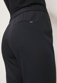 Close-up van een persoon in zwarte elastische broek met een klein achterzakje en een zwart shirt dat erin is gestopt.