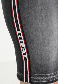 Šedé denimové šortky s bočním pruhem, na kterém je text "REPLAY", lemované červenými a černými pruhy, a s třepeným lemem.