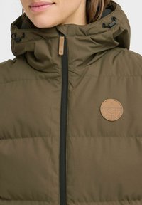 Doudoune vert olive avec un col haut et une capuche ajustable. Présente une fermeture éclair noire et un patch de marque en cuir beige sur la poitrine.