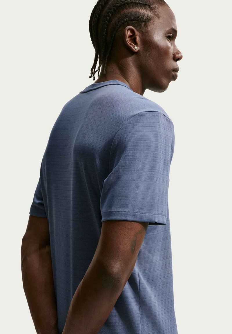 Hombre con cabello trenzado que lleva una camisa azul texturizada de manga corta, mirando hacia la derecha contra un fondo claro y liso.