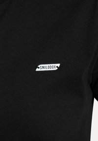 Camiseta negra con textura acanalada que presenta una etiqueta metálica plateada que dice "SMILODOX", cosida en el lado izquierdo.