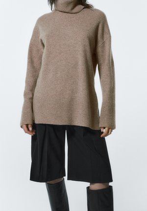 Kobieta ubrana w beżowy, oversize'owy sweter, czarne spodnie culottes oraz czarne kozaki za kolano na jednolitym, jasnym tle.