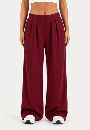 Femme portant un pantalon bordeaux taille haute à jambes larges et des baskets blanches, visible de la taille jusqu'aux pieds sur un fond uni.