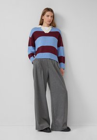 Gestreifter Pullover in Blau und Kastanienbraun, mit V-Ausschnitt und gerippten Bündchen, kombiniert mit grauen Nadelstreifen-Hosen mit weitem Bein und schwarzen Schuhen.