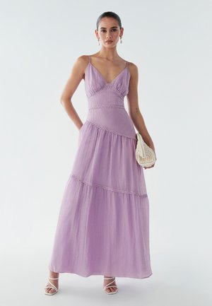 Vrouw in een mouwloze lila maxi-jurk met spaghetti-bandjes, met een witte geweven clutch en witte bandjeshakken.